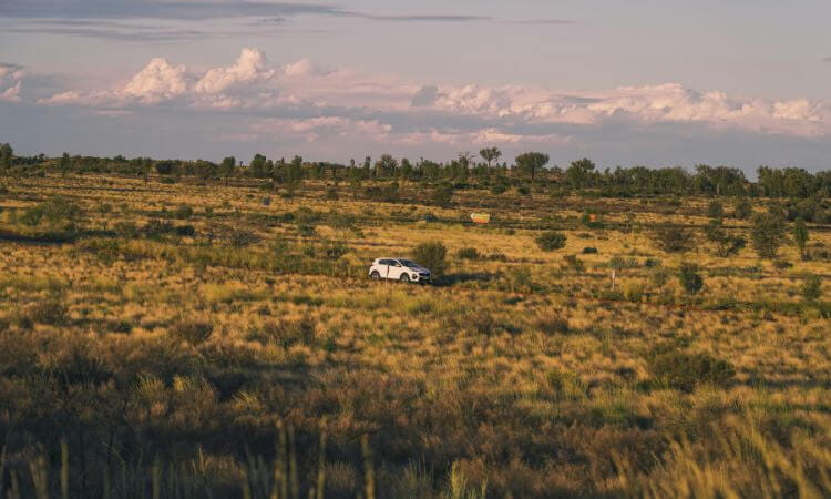 A white car drives through a vast, grassy field under a partly cloudy sky.