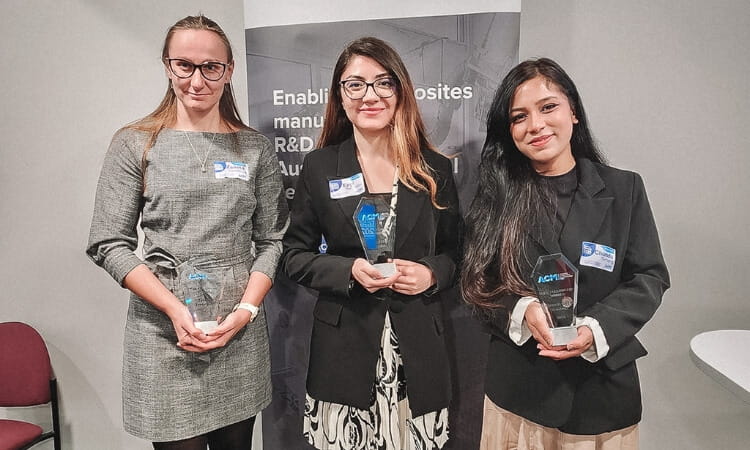 Three women stand smiling, each holding a blue and clear glass award. They are dressed in business attire with a banner backdrop behind them.