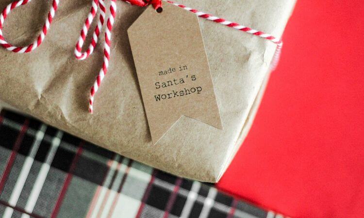 Close-up of a wrapped gift with a tag reading "made in Santa's Workshop." The gift is tied with red and white twine, and part of a plaid-patterned surface is visible underneath.