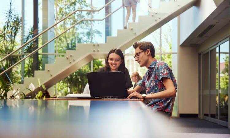 Two people sitting at a table in a modern building, with one using a laptop. There's a large staircase and natural light from windows in the background.