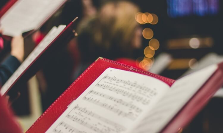 Close-up of red folders holding sheet music, with blurred figures and warm lights in the background.