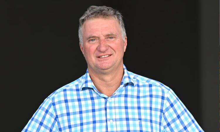 Man in a blue and white checkered shirt smiling against a dark background.