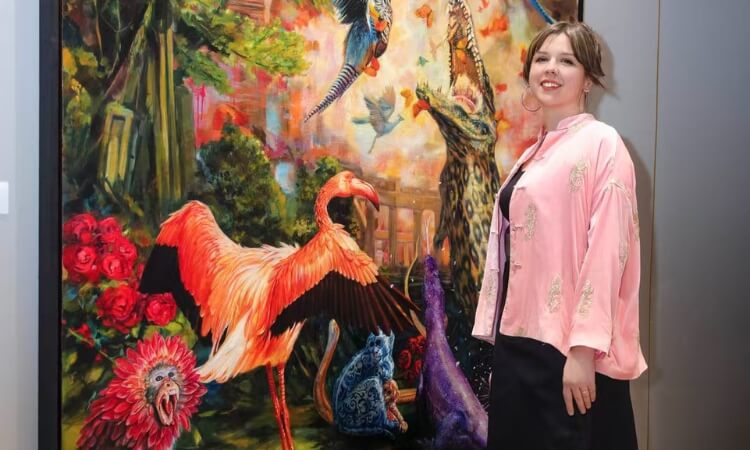 A person stands beside a vibrant painting of various colorful animals, including birds and a flamingo, in a lush, imaginative setting.