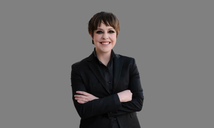 A person with short hair wearing a black suit smiles with arms crossed against a plain gray background.