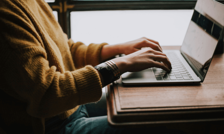 Person in a yellow sweater typing on a laptop at a wooden desk.