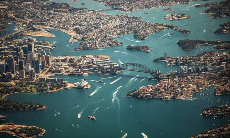 Aerial view of a harbor city with skyscrapers, a prominent bridge, and nearby watercraft activity.