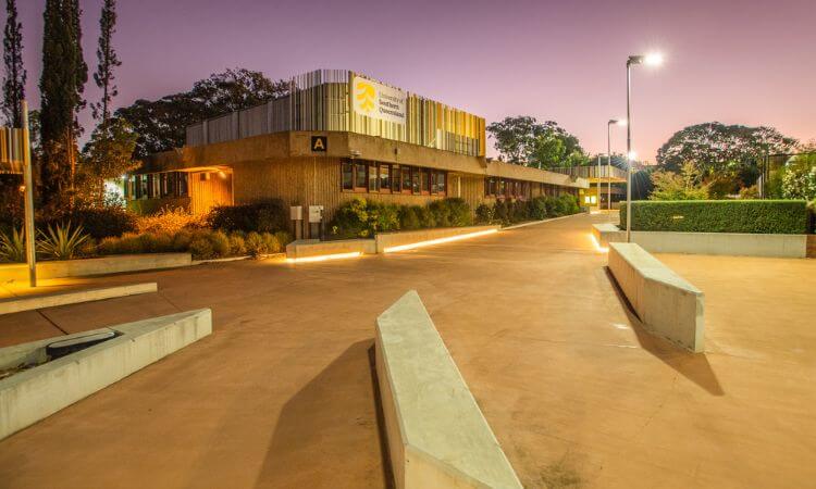 Modern building with a flat roof and large windows, surrounded by lit pathways and greenery, under a pinkish sky at dusk.