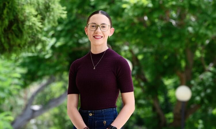 Person wearing glasses and a maroon top smiles outdoors with greenery in the background.