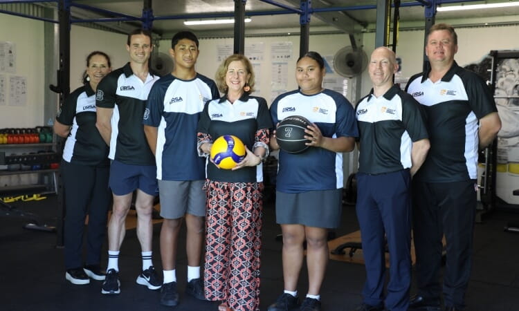 A group of seven people in sports attire stand indoors, with two holding a volleyball and a medicine ball.