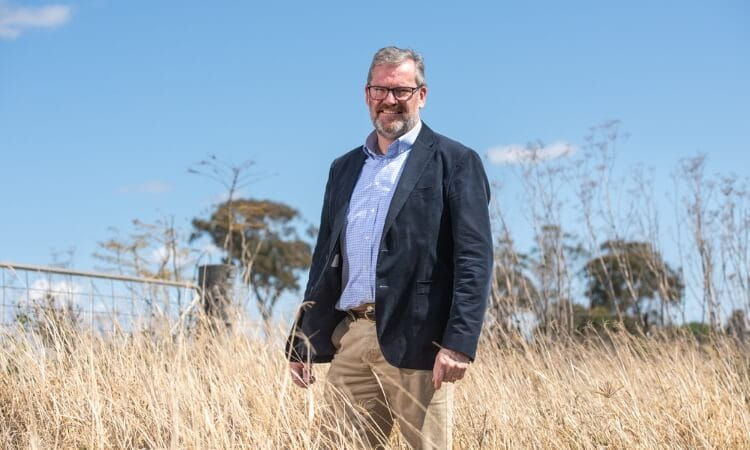 Man in a suit jacket and khakis stands in a dry grassy field under a clear sky.