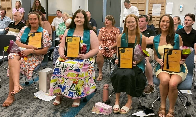 Four women sit in a row, holding certificates and small bouquets, at an indoor ceremony. People are seated and standing in the background, observing the event.