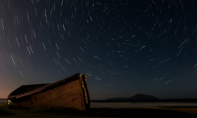 A wooden boat lies on a beach under a night sky with visible star trails, indicating long-exposure photography.