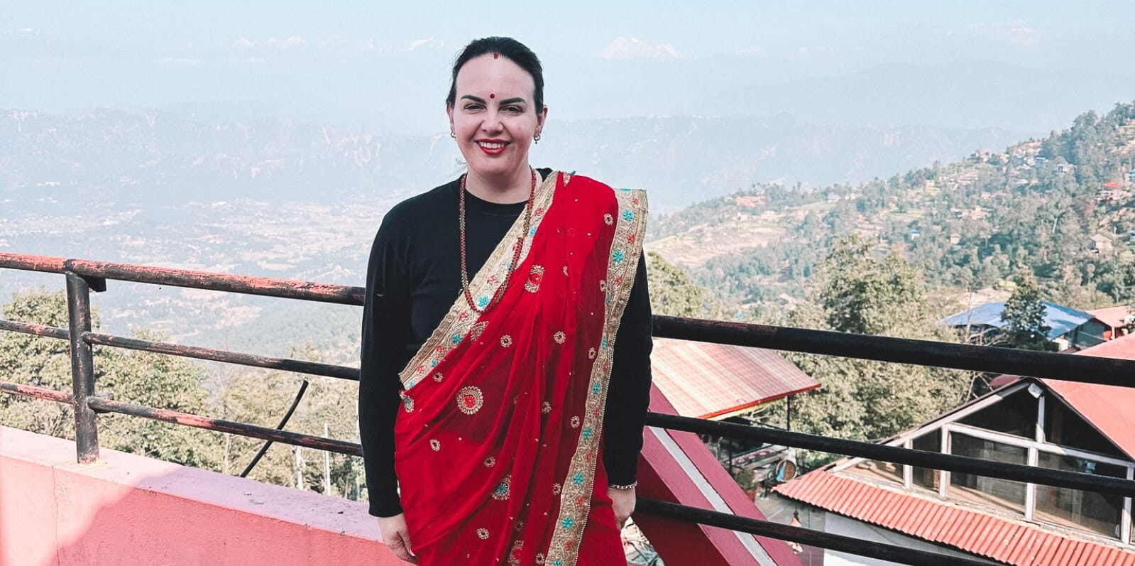 A person in a red sari stands on a terrace overlooking a mountainous landscape with buildings and trees nearby.