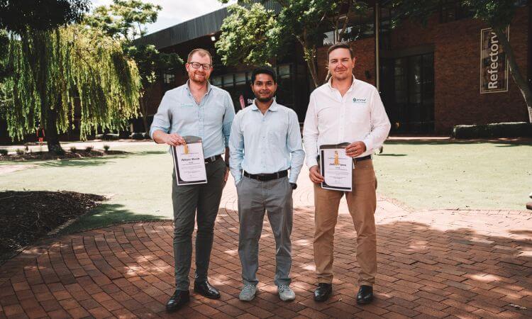 Three men stand outdoors on a brick pathway holding certificates, with trees and a building in the background.