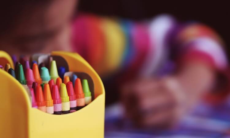 A box of colorful crayons sits in the foreground, while a child wearing a rainbow-striped sleeve colors in the background.