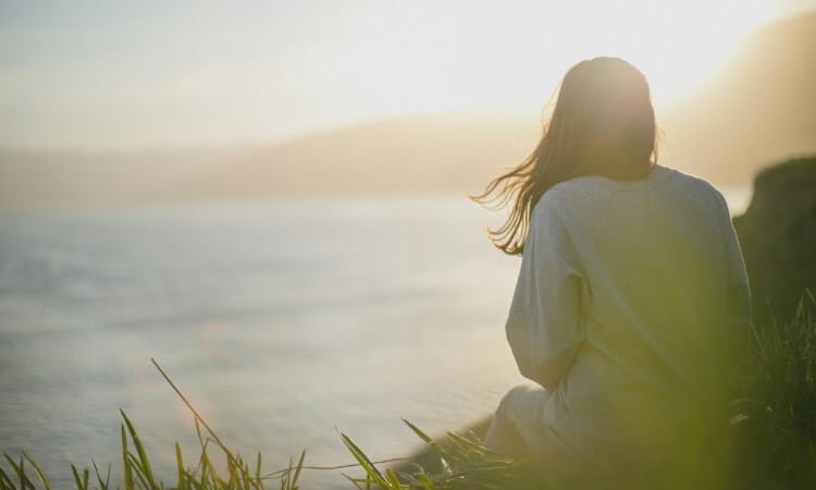 Person sitting on a grassy hill, facing a scenic body of water with sunlight in the background.