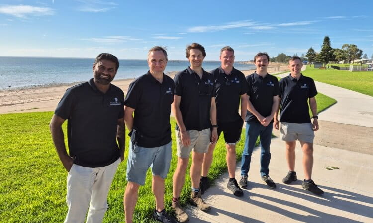 Six people in black shirts stand on a grassy path by the beach, smiling in the sunlight.