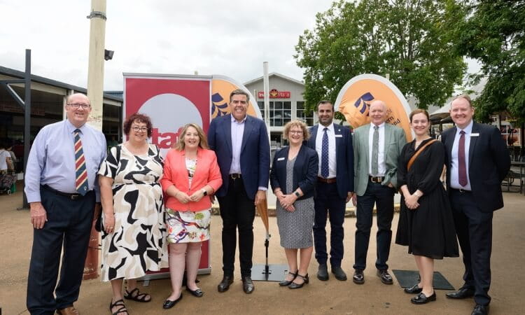 A group of nine people stand outdoors in front of banners, posing for a photo. Some wear suits and others are in business casual attire. Trees and a building are visible in the background.