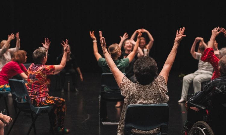 People sitting in chairs, participating in a group activity with raised arms in a dark room.