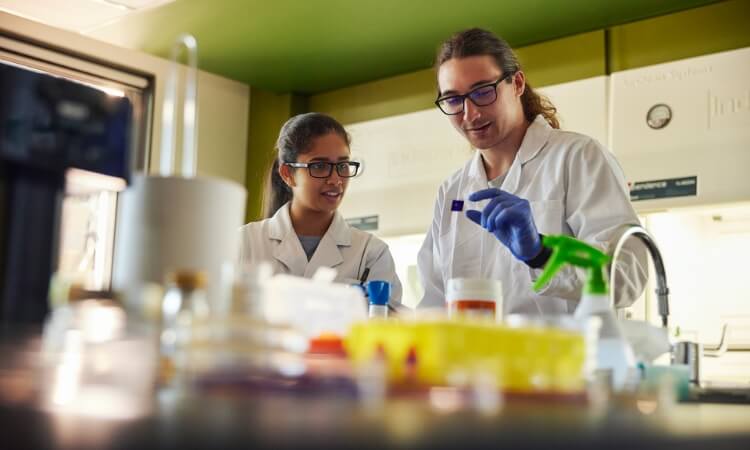 Two scientists in a lab, wearing white coats and safety glasses, examine a slide. Lab equipment and containers are visible in the foreground.