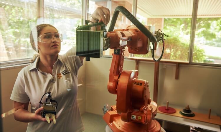 A technician in safety glasses operates an orange robotic arm in a sunny room, using a handheld control device.