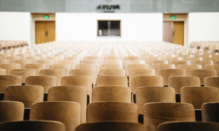 Empty lecture hall with rows of wooden seats facing a stage in a university setting.