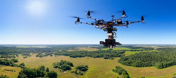 A multi-rotor drone with a camera flies over a vast, green landscape under a clear blue sky.