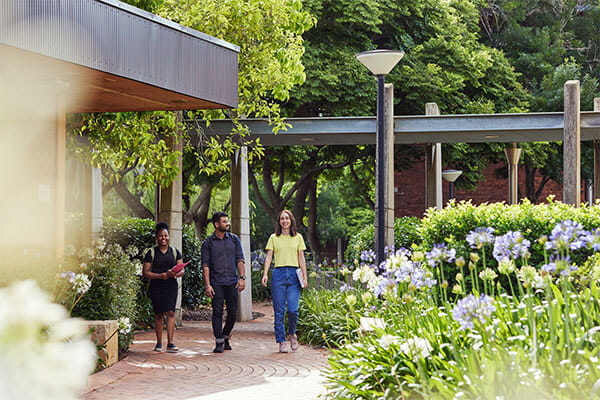 Three people walking along a paved path, surrounded by lush greenery and flowers, with modern buildings in the background.