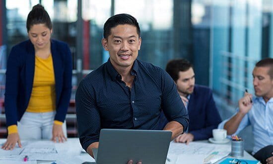 A confident man with a laptop sits at a table in a modern office, with colleagues working in the background.