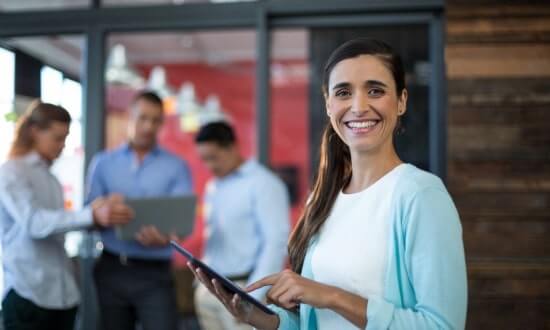 A smiling woman holding a tablet with colleagues working in the background.