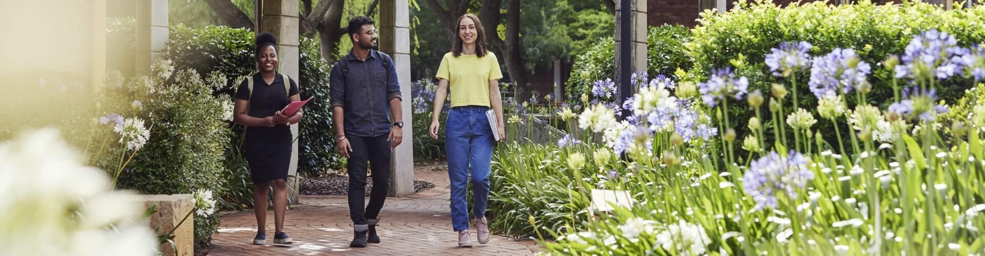 Three people walk along a brick path in a garden with purple flowers and greenery on a sunny day.