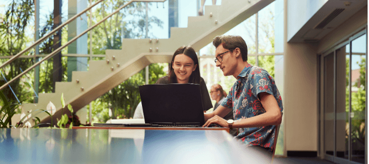 Two people sit at a table working on a laptop in a bright, modern space with large windows and a staircase in the background.