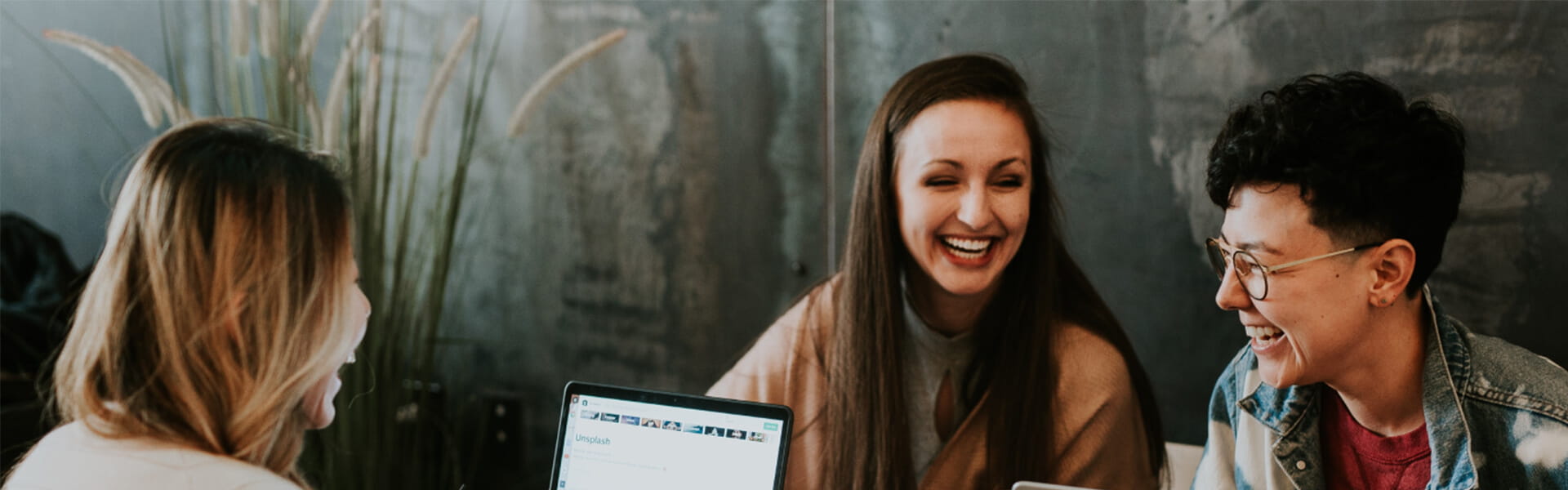 Three people are seated at a table with laptops, smiling and engaging in conversation in a casual setting.