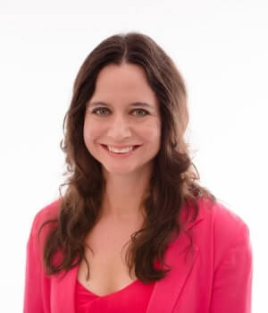 A woman with long brown hair is smiling and wearing a pink blazer over a pink top against a plain white background.