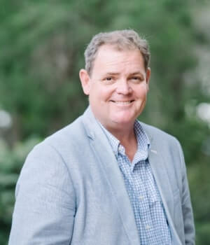 Matt Grant is wearing a light gray blazer and a blue checkered shirt smiles while standing outdoors with greenery in the background.