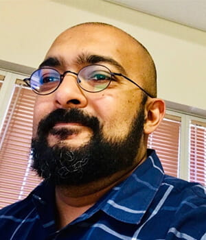 A man with a beard and glasses is wearing a blue striped shirt and standing indoors in front of closed blinds.