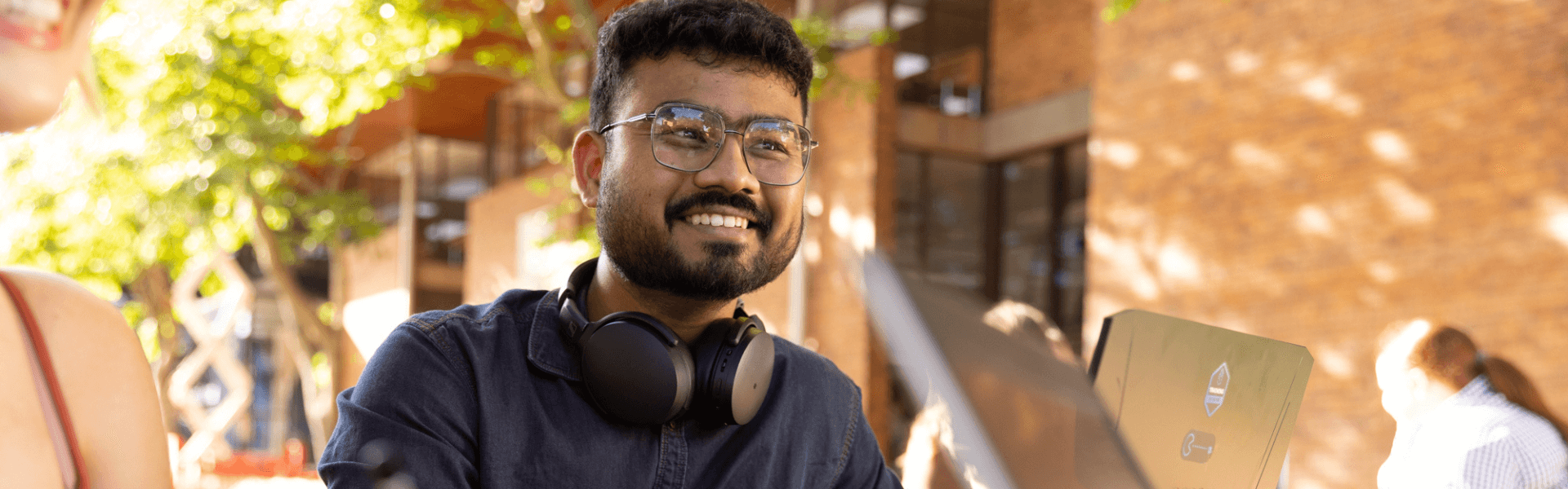 A person wearing glasses and headphones smiles while working on a laptop outdoors in a sunlit area with trees and buildings in the background. 