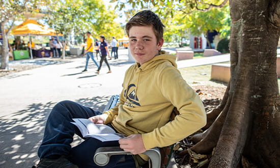 A young person wearing a yellow hoodie sits on a bench under a tree, holding an open book. People walk in the background on a sunny day.