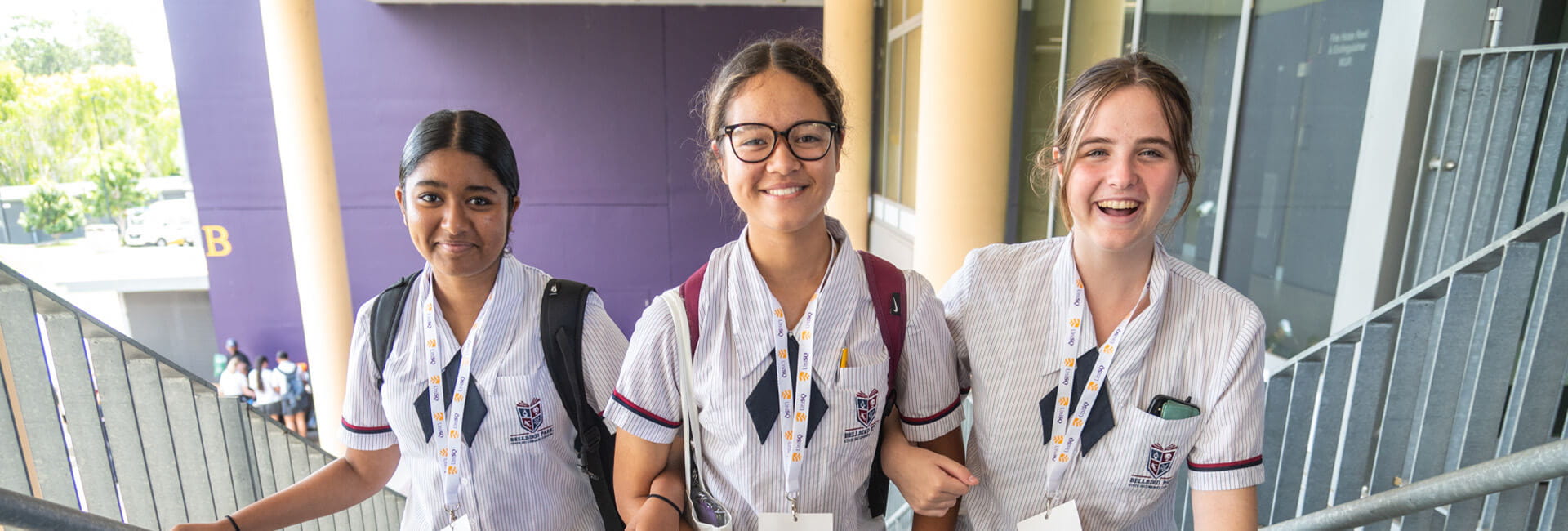 Three students in school uniforms are walking up a staircase, smiling and looking at the camera. 