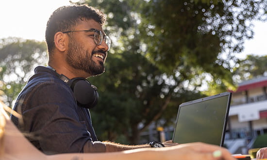 Man with glasses and headphones smiles while working on a laptop outdoors with trees in the background.