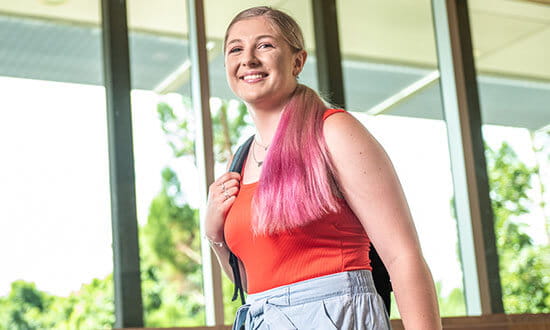 A person with pink hair stands outside, smiling and carrying a backpack.