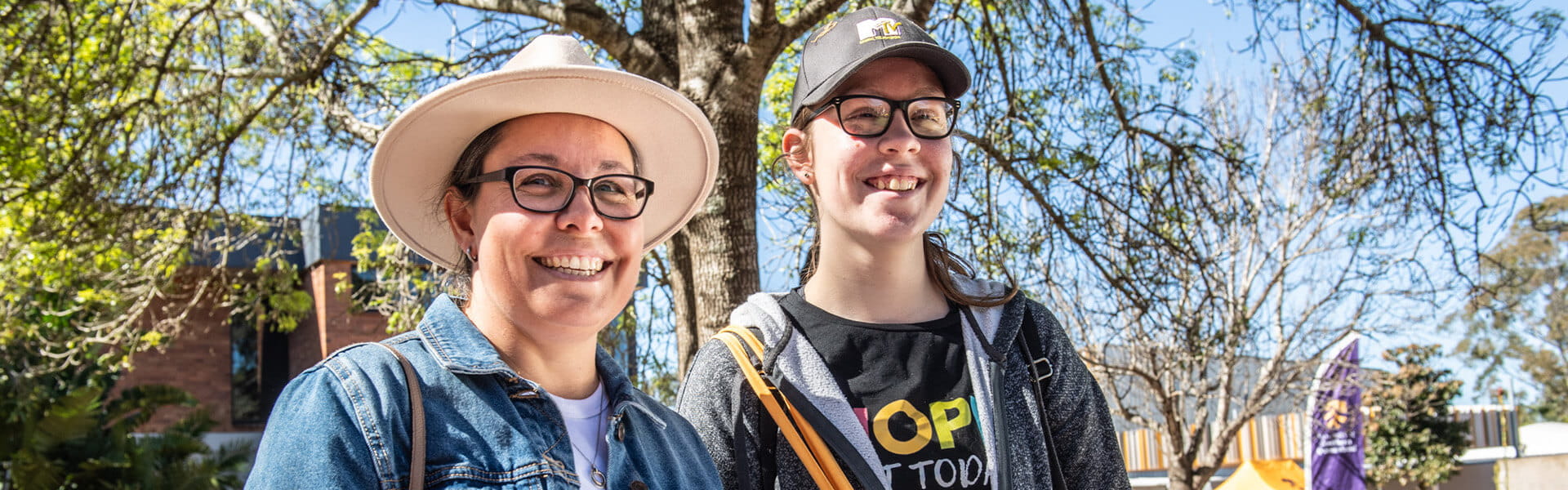 Two individuals smiling outdoors, wearing glasses and hats. Trees and a building are visible in the background on a sunny day.