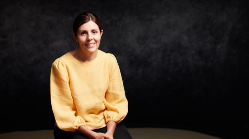 A woman in a yellow blouse and black pants is seated against a dark textured background, smiling gently at the camera.