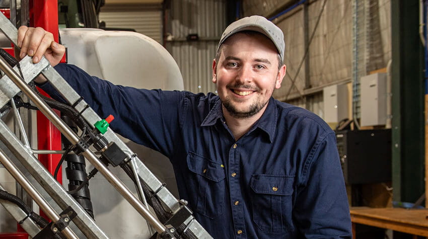 A person wearing a blue shirt and cap is smiling while leaning on agricultural equipment in an industrial setting.