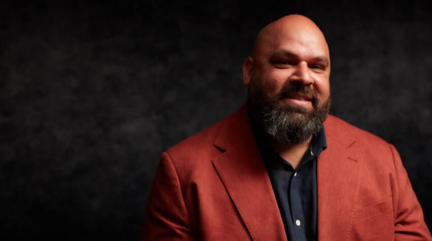 A bearded man in a red blazer and dark shirt is smiling against a dark, textured background.