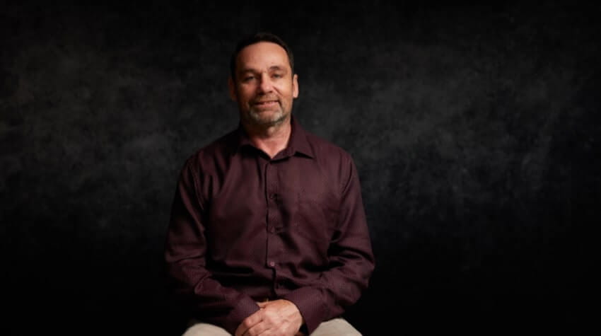 A man with short hair and a beard is sitting and smiling, wearing a dark long-sleeve shirt against a dark background.