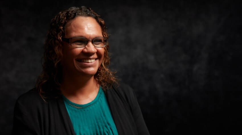 A woman with curly hair and glasses, wearing a green top and dark jacket, is smiling against a dark, textured background.